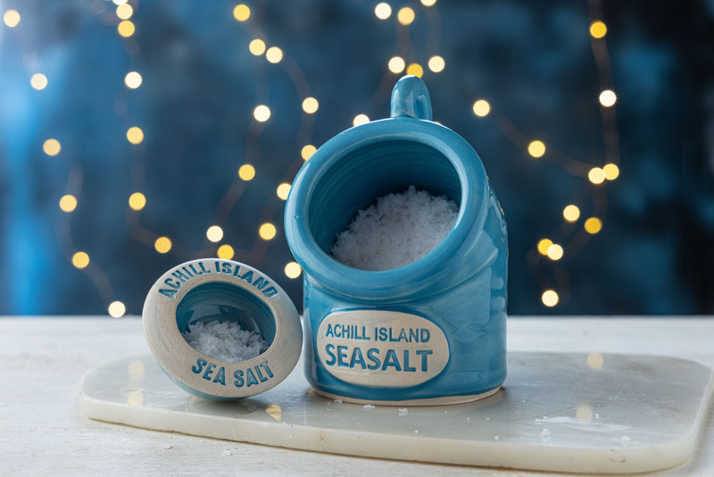 Blue ceramic sea salt pig and pinch pot set with 'Achill Island Seasalt' label on a blurred lights background