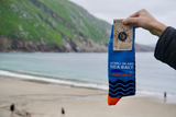 A hand holds a single sock package labeled Achill Island Sea Salt Pure wild against a keem bay background.