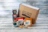Achill Island Sea Salt gift box set on a light blue wooden surface, featuring a large brown cardboard box, a blue ceramic bowl, a wooden spoon, and packaged sea salt and crackers.