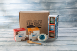 Achill Island Sea Salt gift set arranged on a light blue wooden surface, including a large brown branded box, a red and white nougat box box, an orange and white fudge box, a tall blue-and-white package, a small blue-and-white bowl, and a wooden spoon.