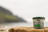 A small glass jar of Achill Island Sea Salt (Wakame Seaweed) with a dark green lid sits on a wet rock in the foreground, with a blurred coastal landscape in the background.