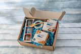 An open cardboard gift box filled with blue crinkle paper and assorted Achill Island Sea Salt products, including jars, a wooden spoon, and packaged snacks, photographed from above on a light blue wood plank surface.
