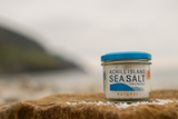A small glass jar of Achill Island Sea Salt with a blue lid sits on a rock outdoors with a blurred coastal background.