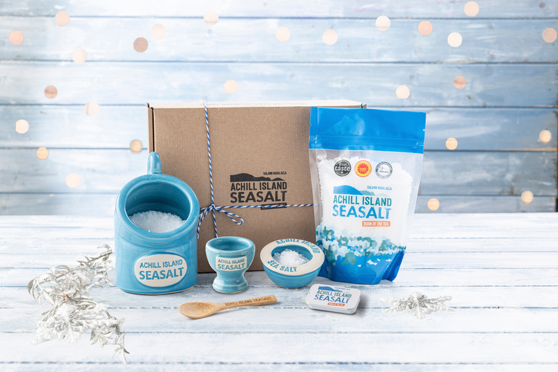 Achill Island Seasalt products including a salt shaker, packets, and containers on a wooden surface with a light blue background.