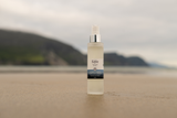 A bottle of Säle Sea Salt body oil standing on a sandy beach with a blurred coastal landscape in the background.