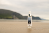 A bottle of Saile Facial Spritz standing on a sandy beach with a blurred mountain background.