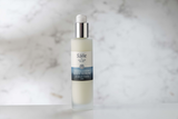 Säile Body Serum in a frosted glass bottle with a white pump dispenser, set against a soft, blurred white marble background.
